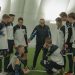A soccer coach gives tactical advice to a youth team indoors on artificial turf.